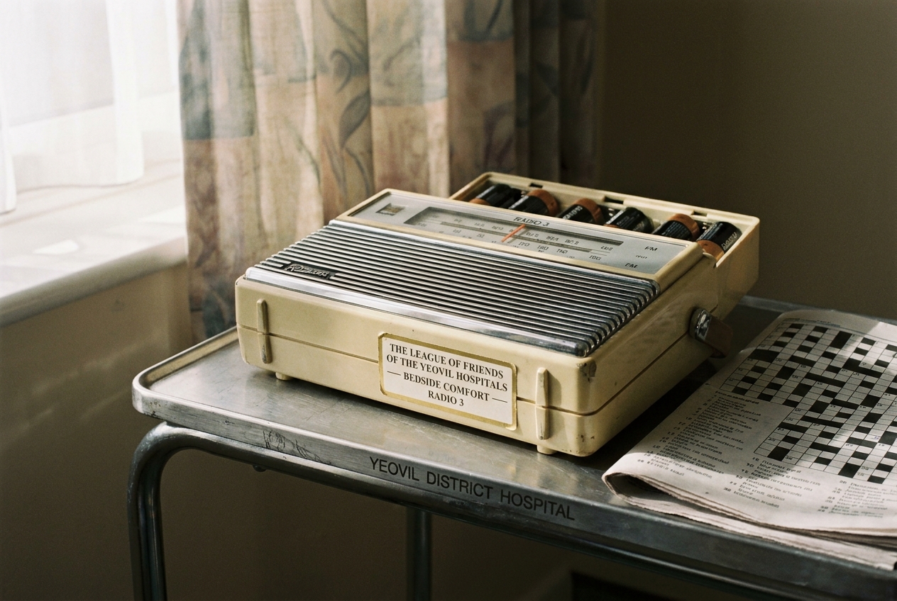 A small Roberts bedside radio on a Yeovil District Hospital tray-table, tuned to Radio 3, the cream-and-gold Friends&rsquo; sticker on the back of the casing reading &lsquo;THE LEAGUE OF FRIENDS OF THE YEOVIL HOSPITALS&rsquo;.